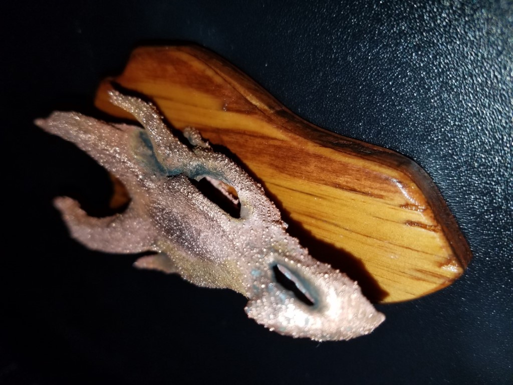 Top-down close-up of a silvery sintered-metal fragment with a porous beaded texture and bluish-green patina on an amber wooden paddle-shaped base