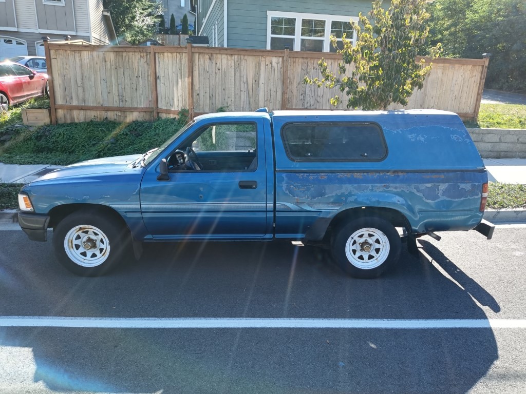 Weathered blue single-cab pickup with peeling paint revealing rust on the bed and shell, parked before a wooden privacy fence