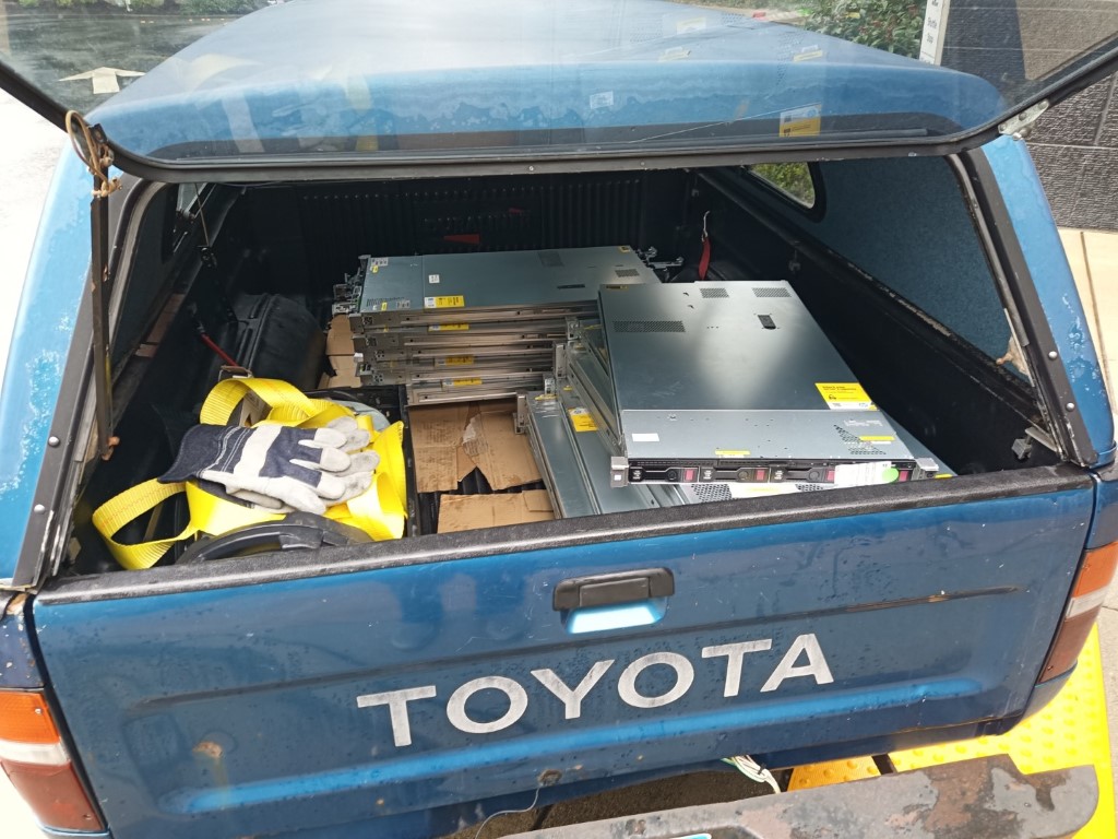 Rear of the blue Toyota pickup with the bed loaded with rack-mount servers secured by yellow ratchet straps