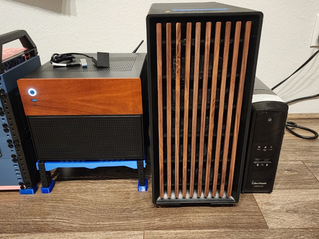 Small wood-topped PC on a blue 3D-printed stand, a wood-slat front tower, and a CyberPower UPS on a laminate floor