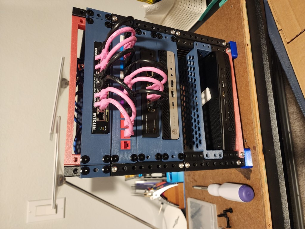 3D-printed mini server rack holding a network switch, an HP mini PC, and a mounted chassis with coiled pink ethernet cables