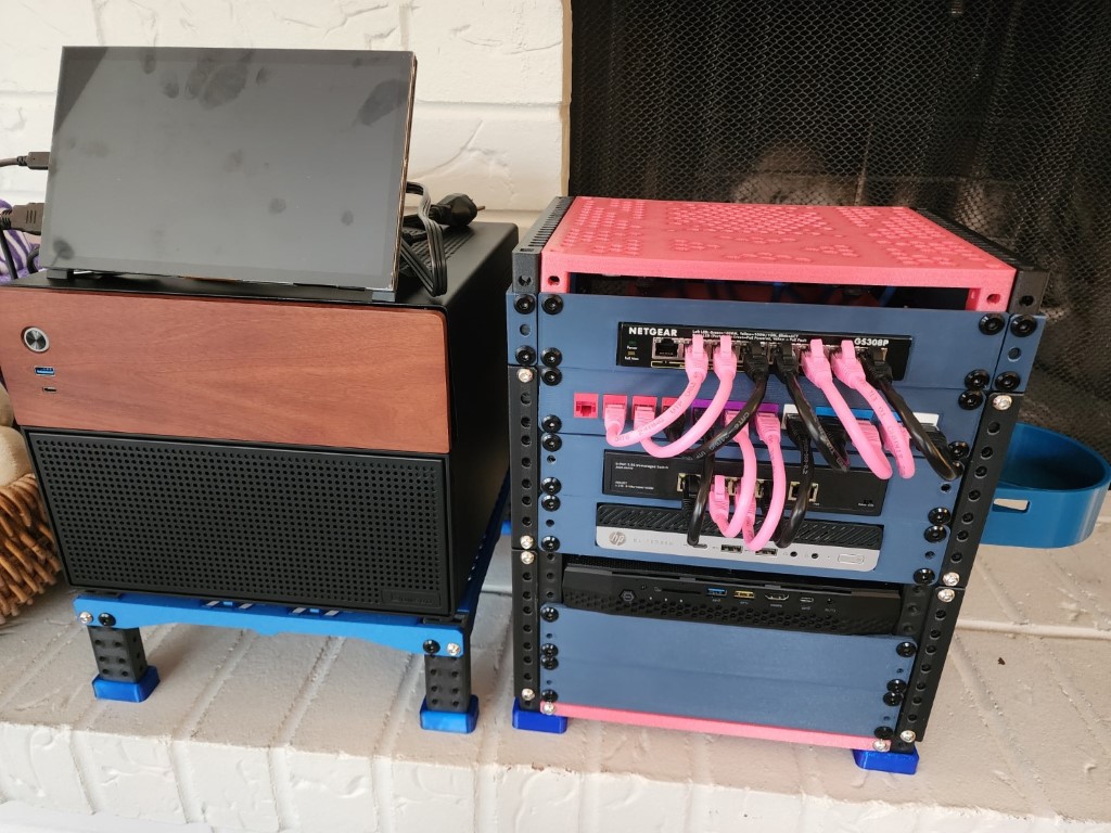Home server on a white brick hearth: a black wood-front tower with monitor and a blue rack of networking gear with pink cables