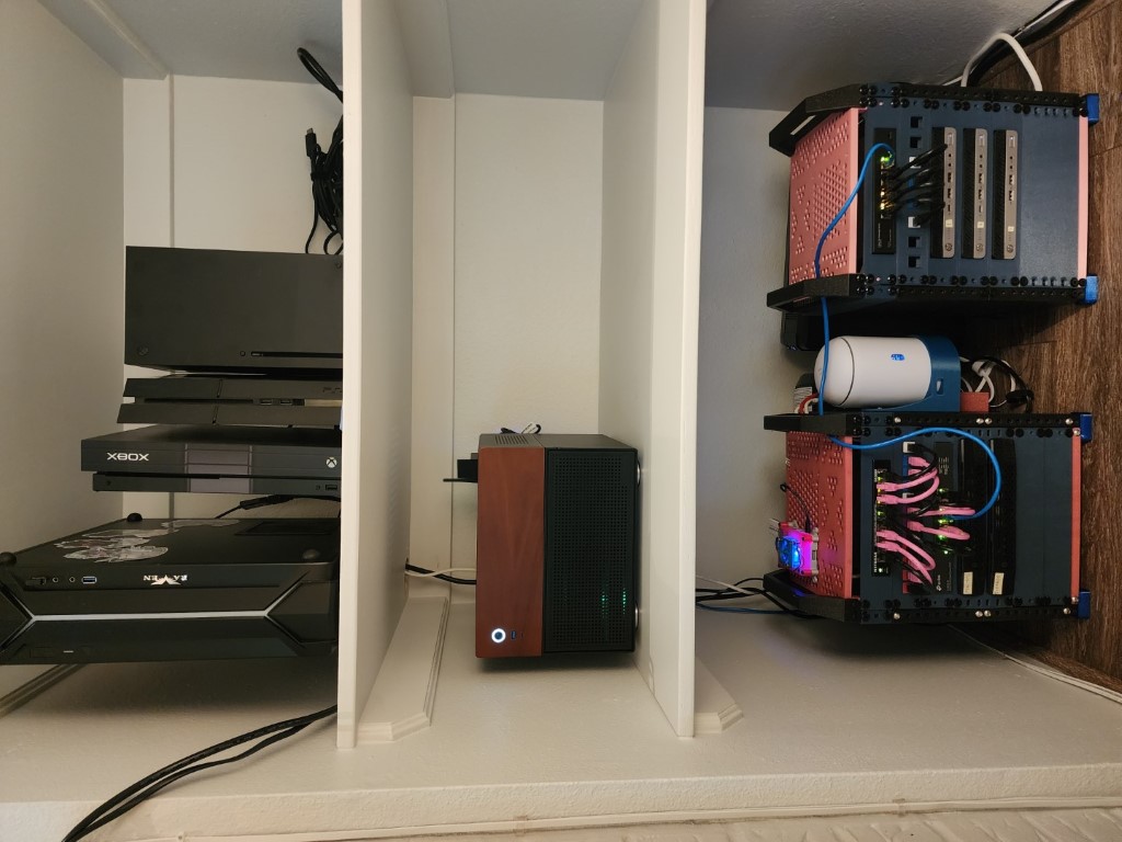 Built-in shelving with gaming consoles, a wood-paneled SFF PC, and a custom server rack with pink side panels and pink and blue cables