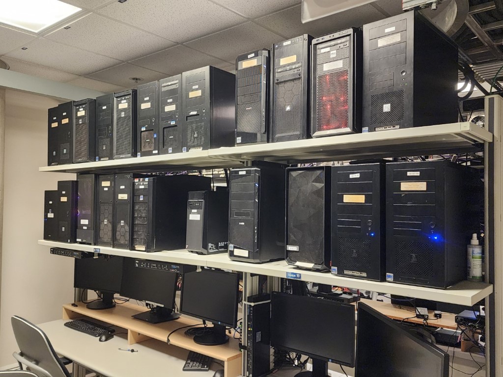 Computer lab storage room with shelves stacked with black desktop towers and desks labeled Station 19